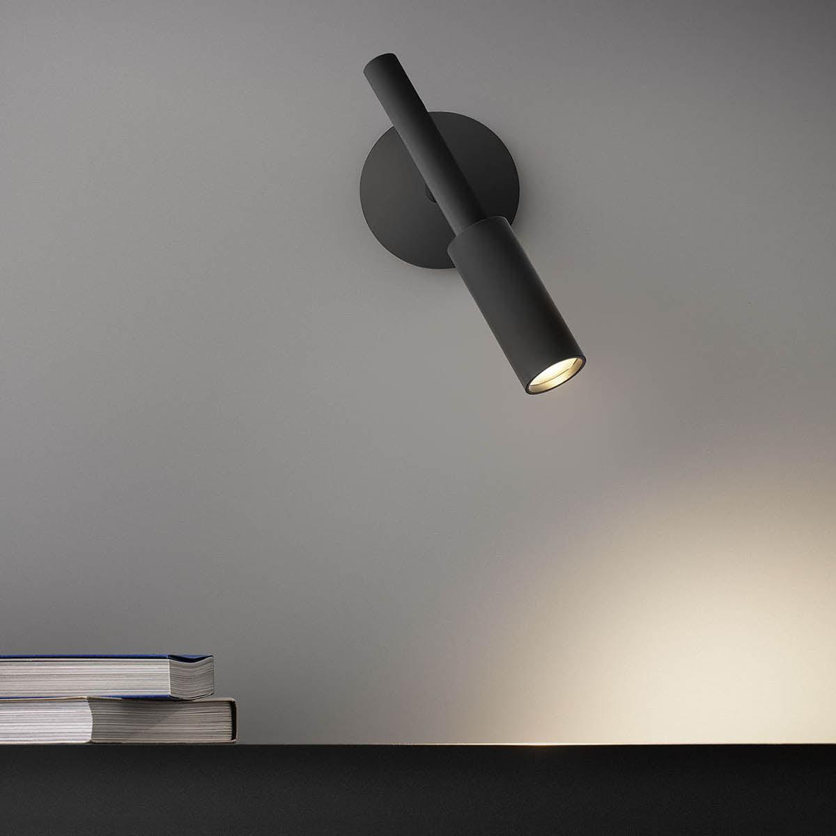 Black Clérachic wall light tilted toward the shelf below, its warm beam falling across the grey wall and a stack of books.