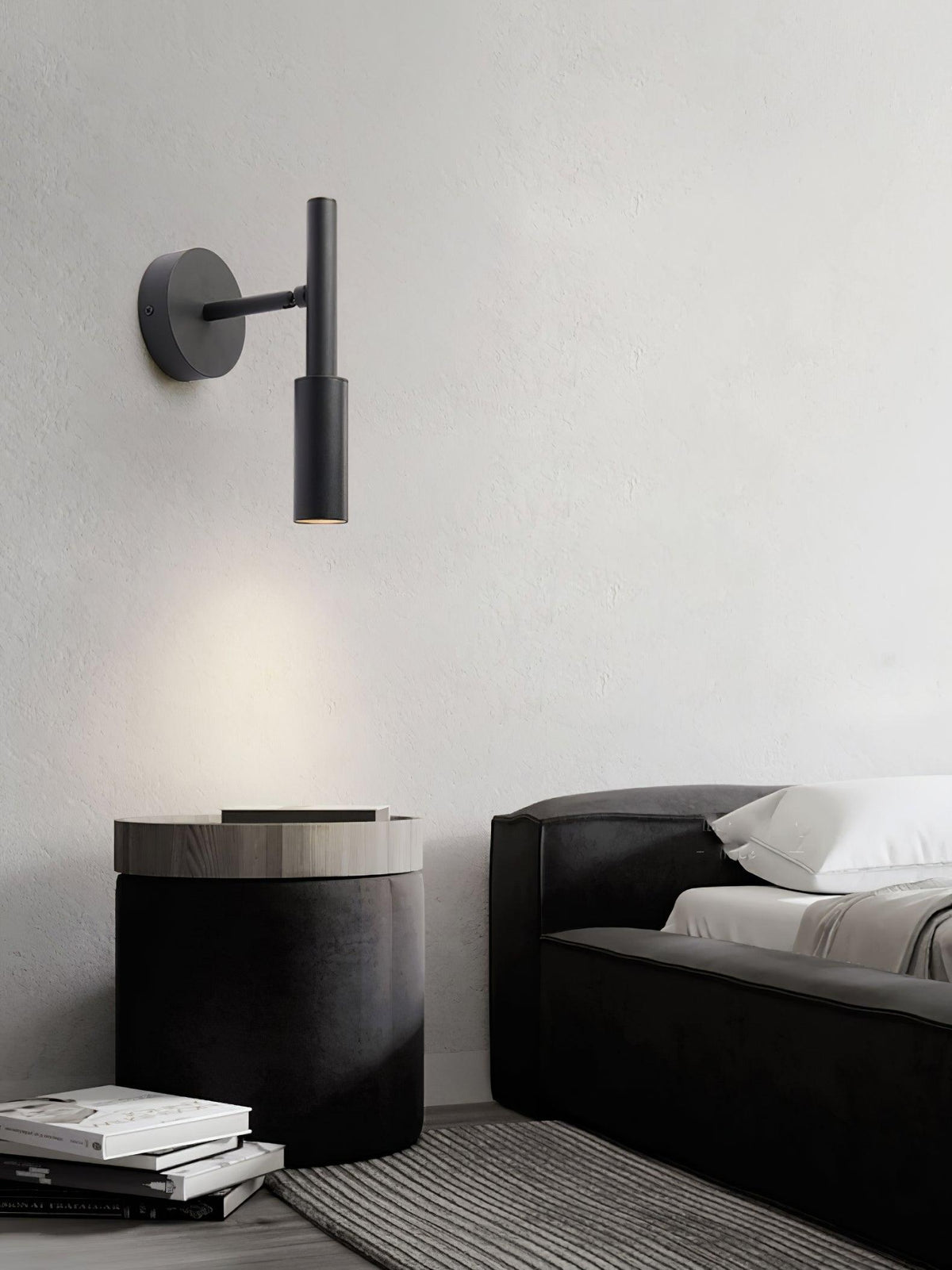 Black Clérachic wall light on a pale wall above a round black pouffe and a low sofa, its beam pointing down.
