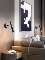 Two black Clérachic wall lights on either side of an abstract print above a tan leather sofa and a dark side table.