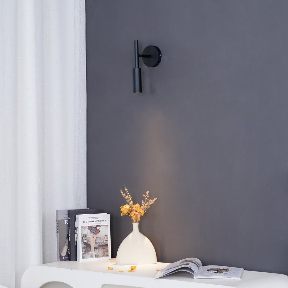 Black Clérachic wall light on a slate-grey wall beside a white curtain, with a soft beam above a console and a vase of dried flowers.