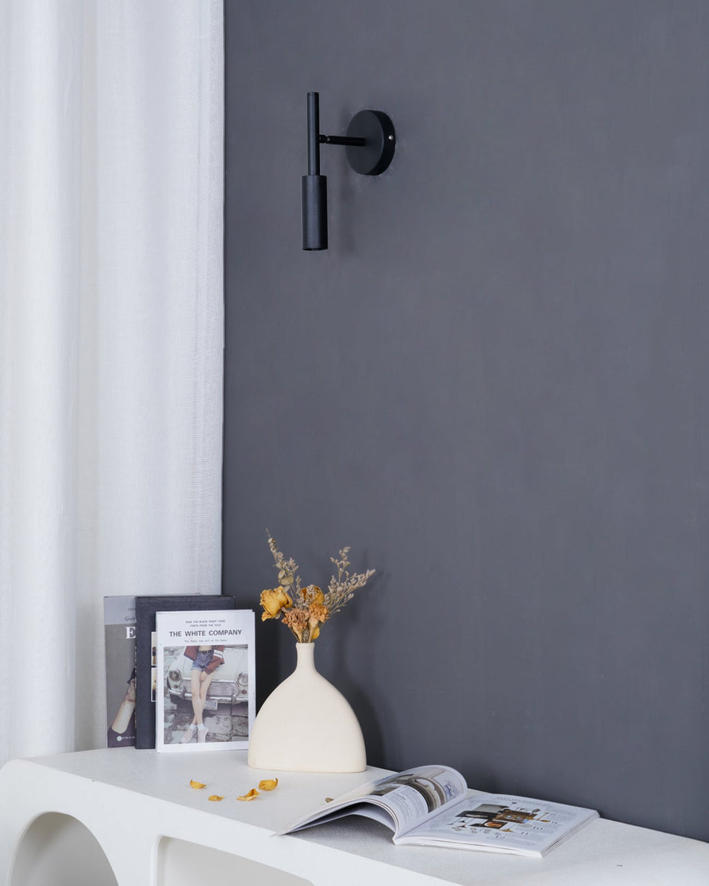 A slim black Clérachic wall light on a dark grey wall, above a white console with a book, flowers and a vase.