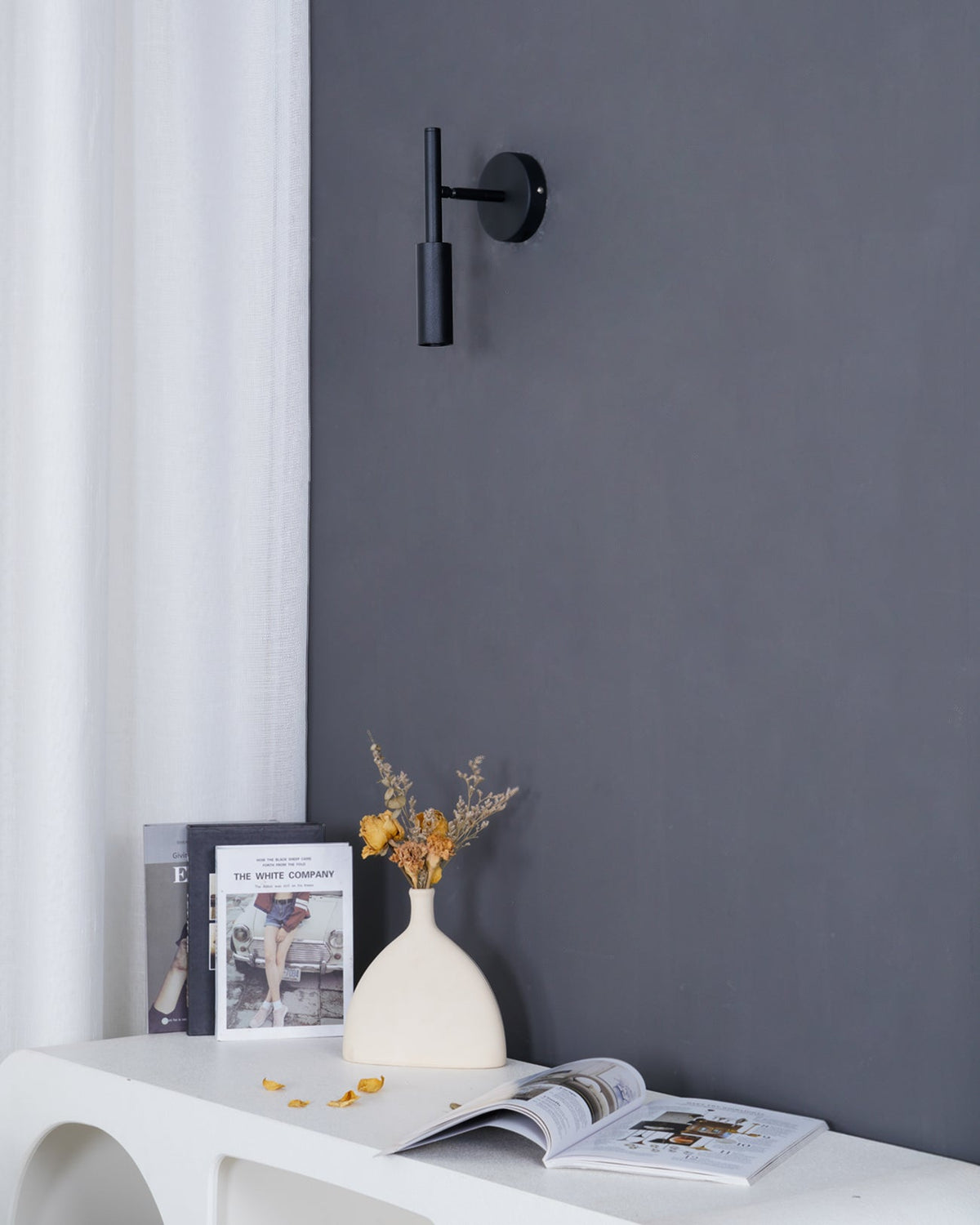 A slim black Clérachic wall light on a dark grey wall, above a white console with a book, flowers and a vase.