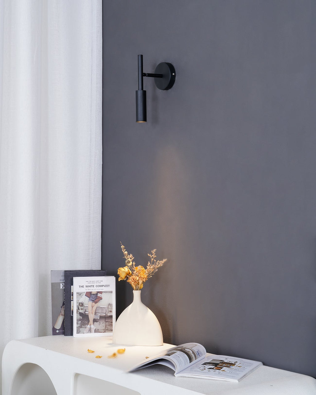 Black Clérachic wall light on a dark grey wall by a white curtain, with its beam falling on a console and a vase of flowers.