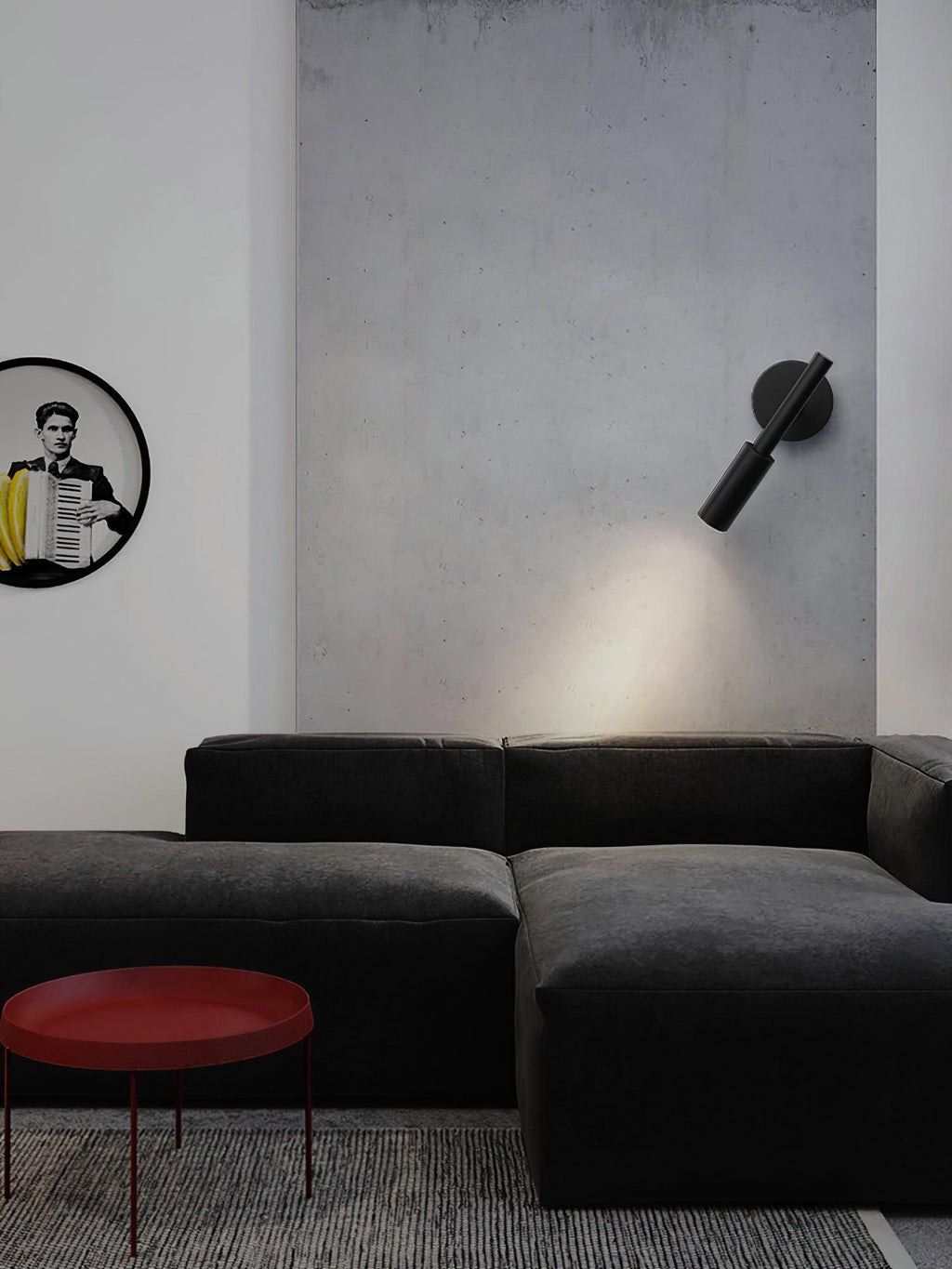 A black Clérachic wall light angled down on a grey concrete wall above a dark sofa and a red round side table.