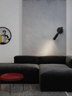 A black Clérachic wall light angled down on a grey concrete wall above a dark sofa and a red round side table.