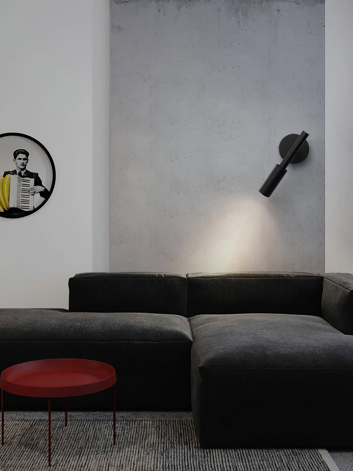 A black Clérachic wall light angled down on a grey concrete wall above a dark sofa and a red round side table.