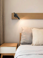 Black Clérachic wall light on a wooden shelf above a white bed, with its warm beam falling on the pillows beside a nightstand.