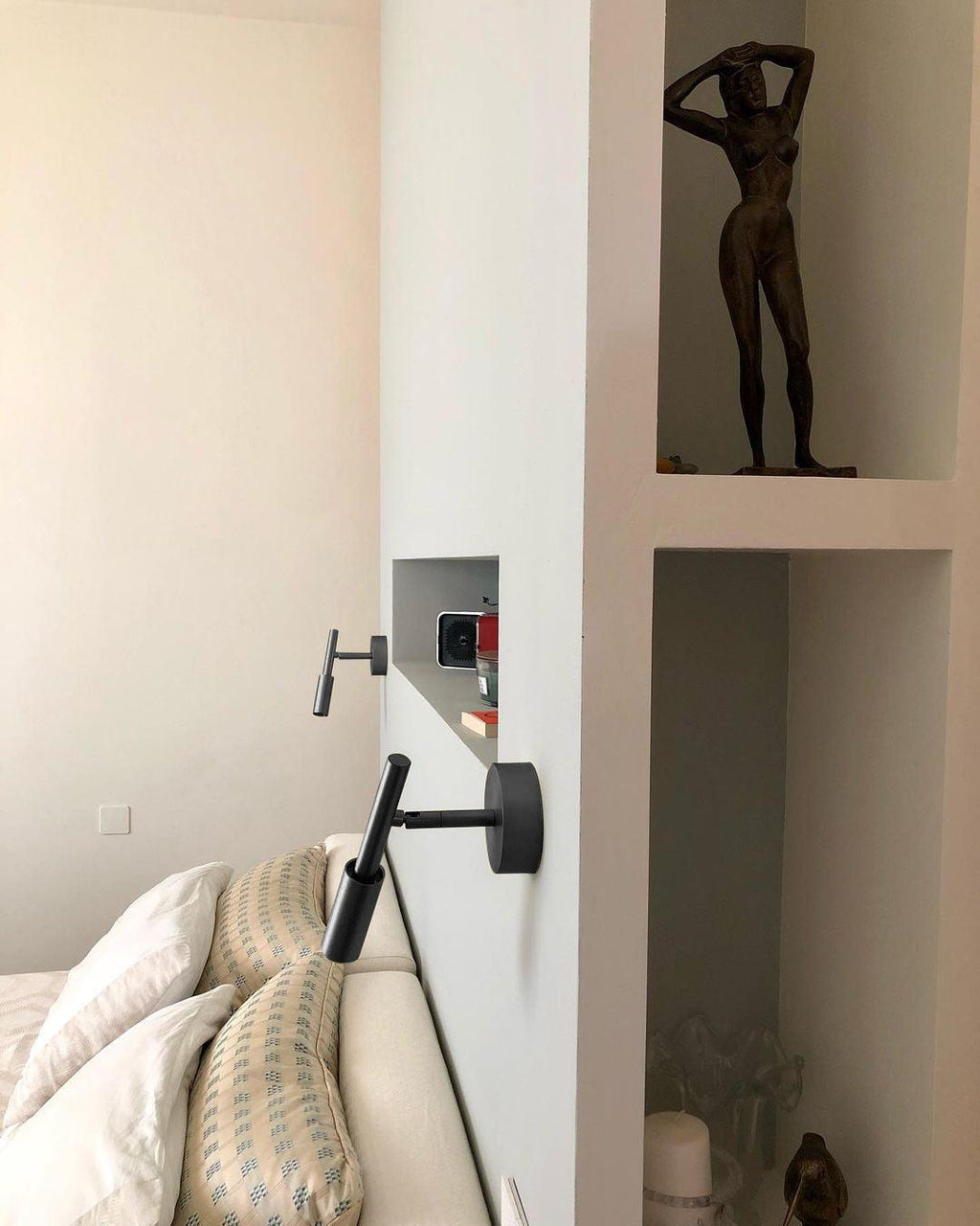 A black Clérachic wall light swung out from the wall beside a bed, with a wall niche and a sculpture behind it.