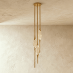 Clérachic three-head pendant in brushed gold with slim rods and clear glass teardrops, hung from a round canopy on a plaster wall.