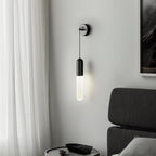 Black Clérachic wall light beside a sheer white curtain and a dark framed picture, with a charcoal bed headboard and a round bedside table.