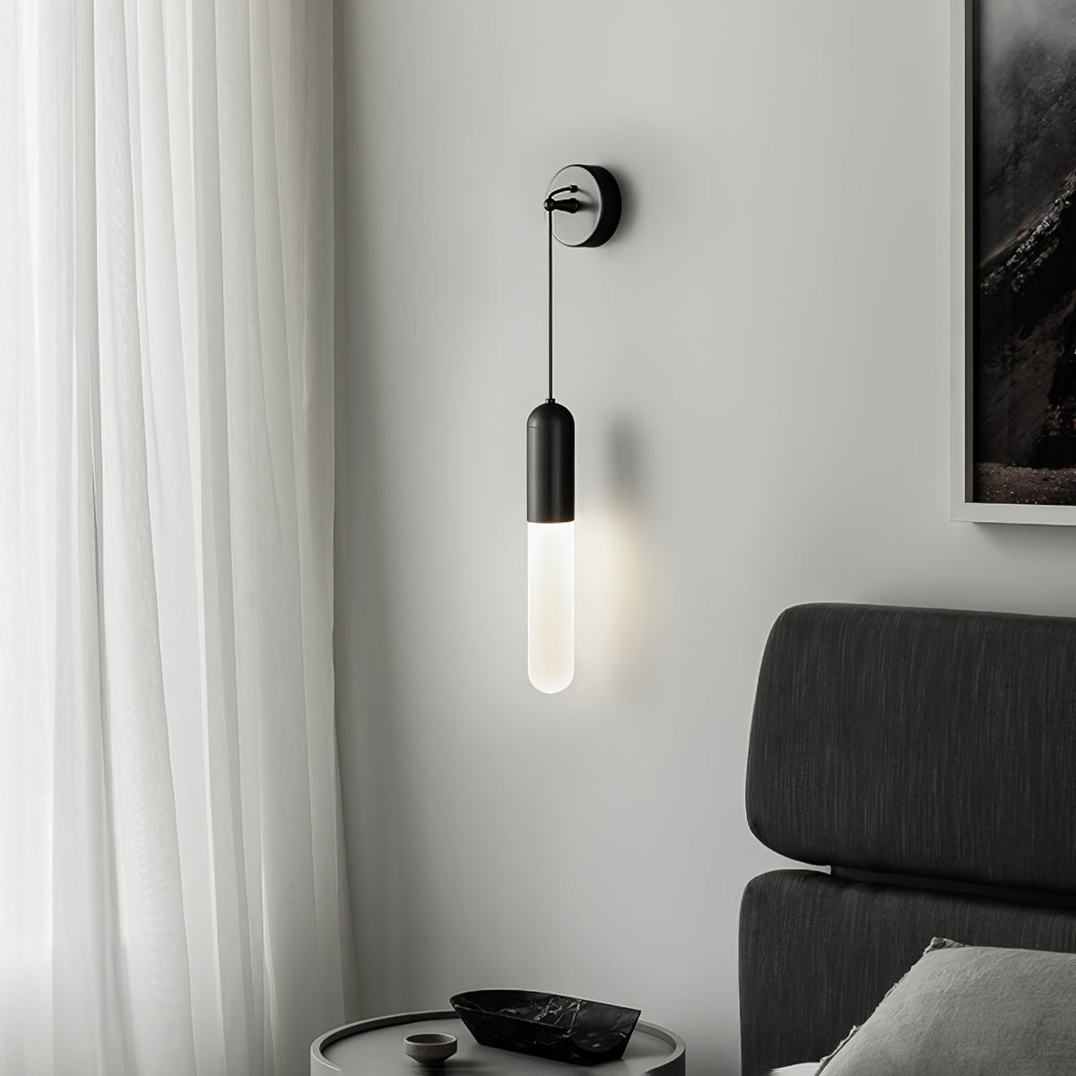 Black Clérachic wall light beside a sheer white curtain and a dark framed picture, with a charcoal bed headboard and a round bedside table.