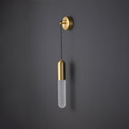 Unlit brass Clérachic wall light on a dark grey wall, the slim frosted cylinder shade pale and the brass cap and wall plate catching the light.