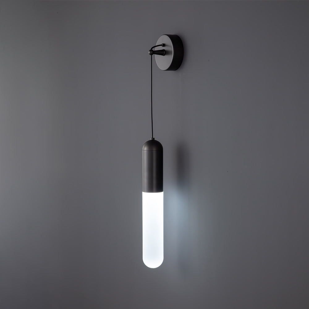 Black Clérachic wall light lit in a cool white light on a grey wall, the slim cylinder shade glowing bright below its dark cap.