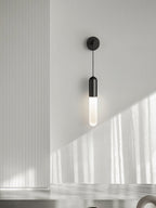 Black Clérachic wall light on a pale grey wall with soft window light, a vertical panel to the left and the shade glowing warm.