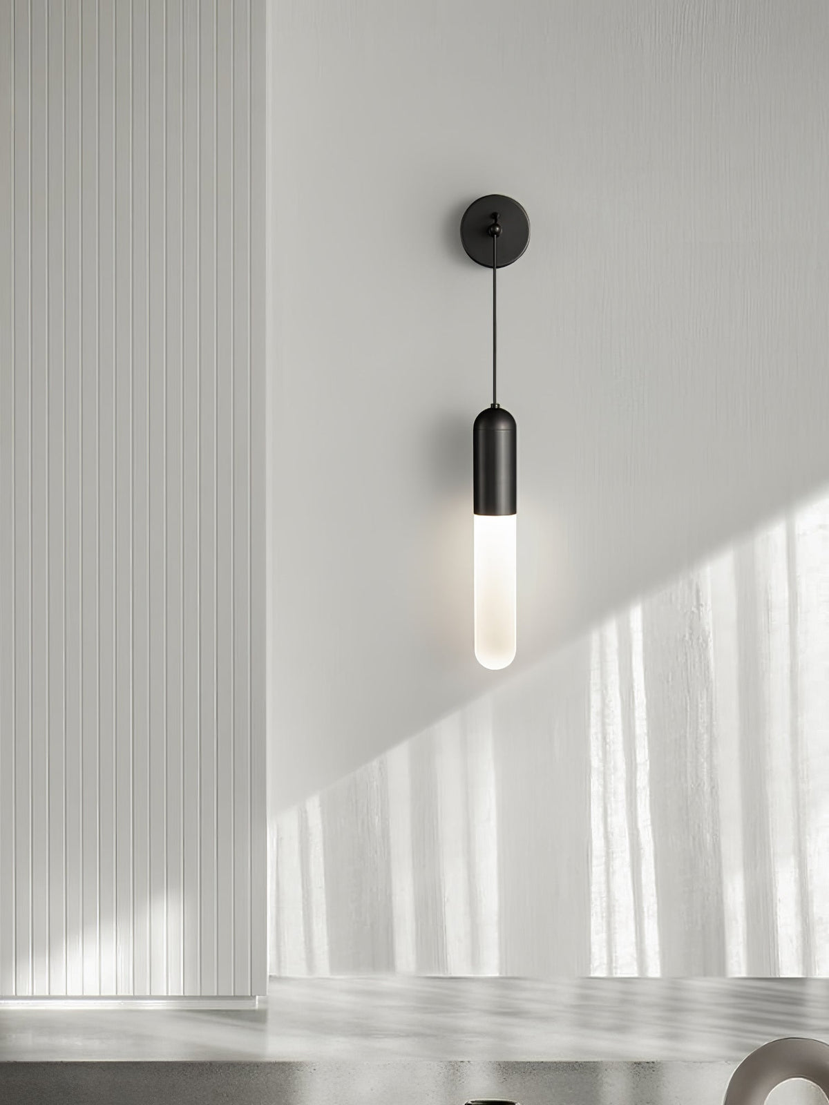 Black Clérachic wall light on a pale grey wall with soft window light, a vertical panel to the left and the shade glowing warm.