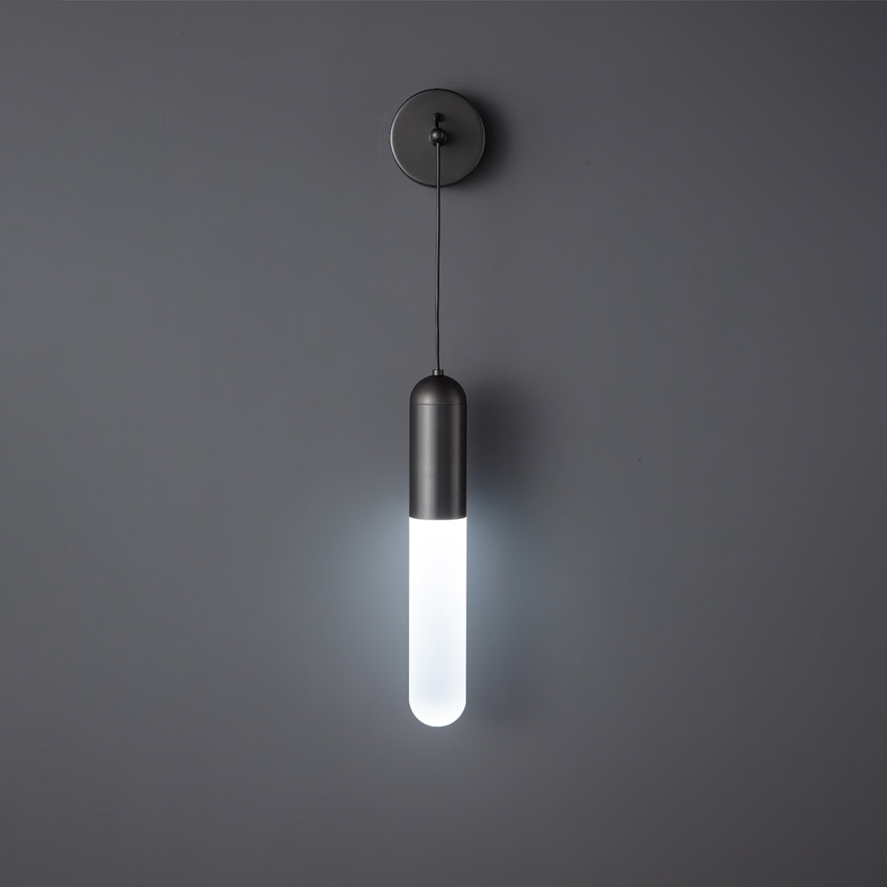 Black Clérachic wall light on a dark grey wall, the slim cylinder shade lit in a cool white light below its black cap.