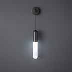 Black Clérachic wall light on a dark grey wall, the slim cylinder shade lit in a cool white light below its black cap.
