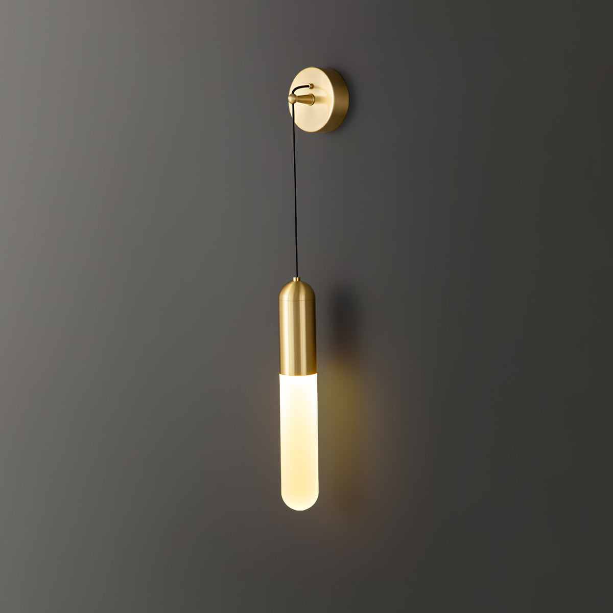 Brass Clérachic wall light on a dark wall, the slim cylinder shade lit warm and casting a soft amber glow on the wall beside it.