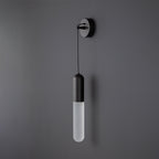 Unlit black Clérachic wall light on a dark grey wall, the frosted cylinder shade pale beneath a black cap and wall plate.