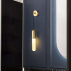 Brass Clérachic wall light on a navy panelled wall beside a dark glass door, its slim shade lit warm and a curved mirror glowing nearby.
