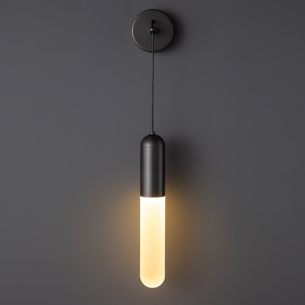 Black Clérachic wall light lit warm on a charcoal wall, the glow spreading softly below the black cap from its frosted cylinder shade.