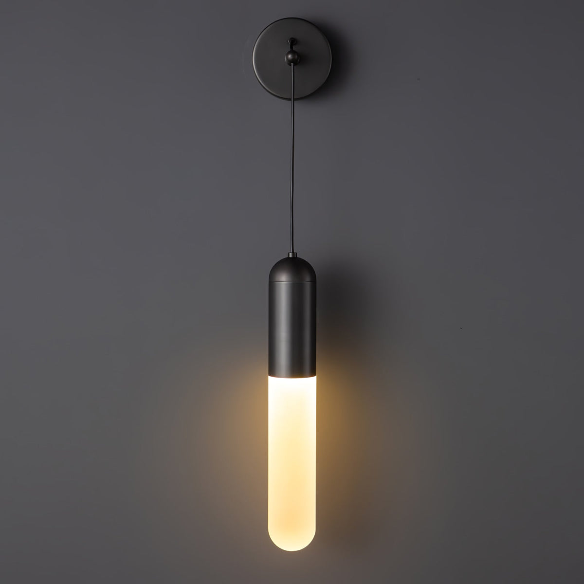 Black Clérachic wall light lit warm on a charcoal wall, the glow spreading softly below the black cap from its frosted cylinder shade.