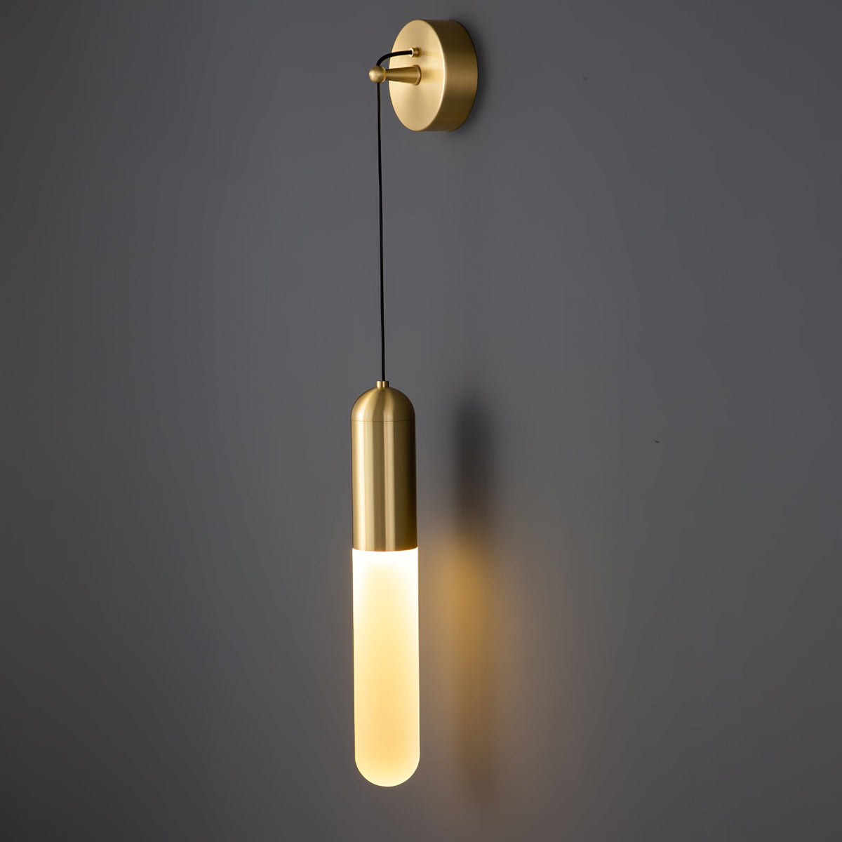 Brass Clérachic wall light on a dark grey wall, the cylinder shade glowing warm and casting a faint shadow to the right.