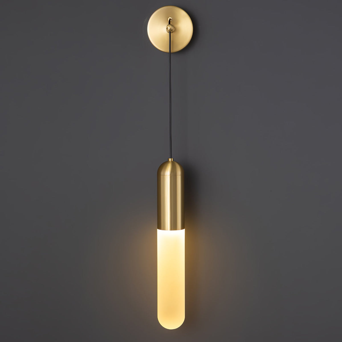 Brass Clérachic wall light up close on a dark grey wall, the round brass plate above a lit cylinder shade with a bright warm glow.