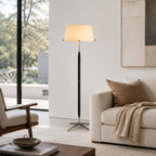 A Clérachic floor lamp stands beside a cream sofa with a rust cushion and a knitted throw, in front of a large window and a framed print.