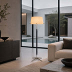 A Clérachic floor lamp with a cream shade stands in front of a glass wall with a courtyard pool, beside a cream sofa and a dark console.