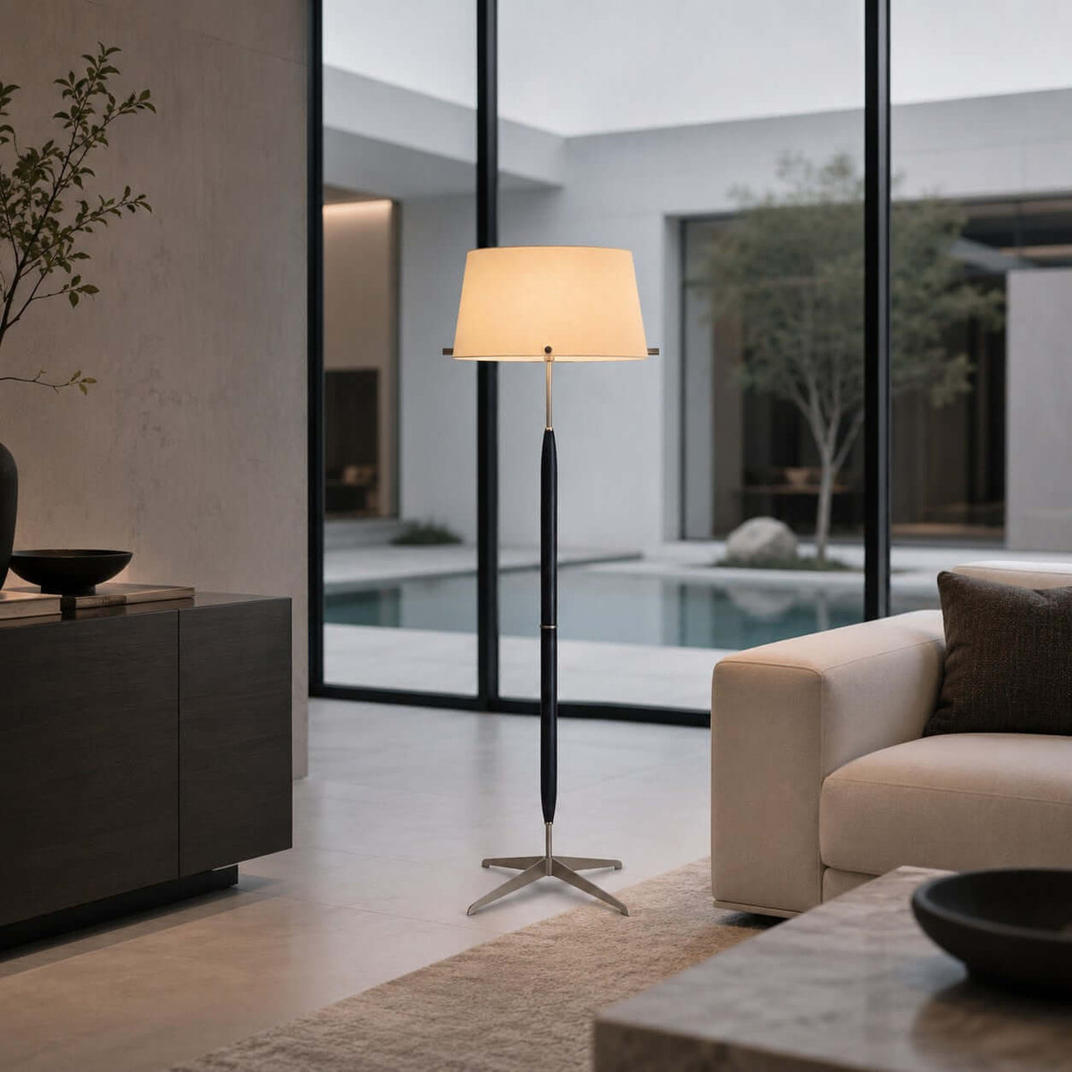 A Clérachic floor lamp with a cream shade stands in front of a glass wall with a courtyard pool, beside a cream sofa and a dark console.