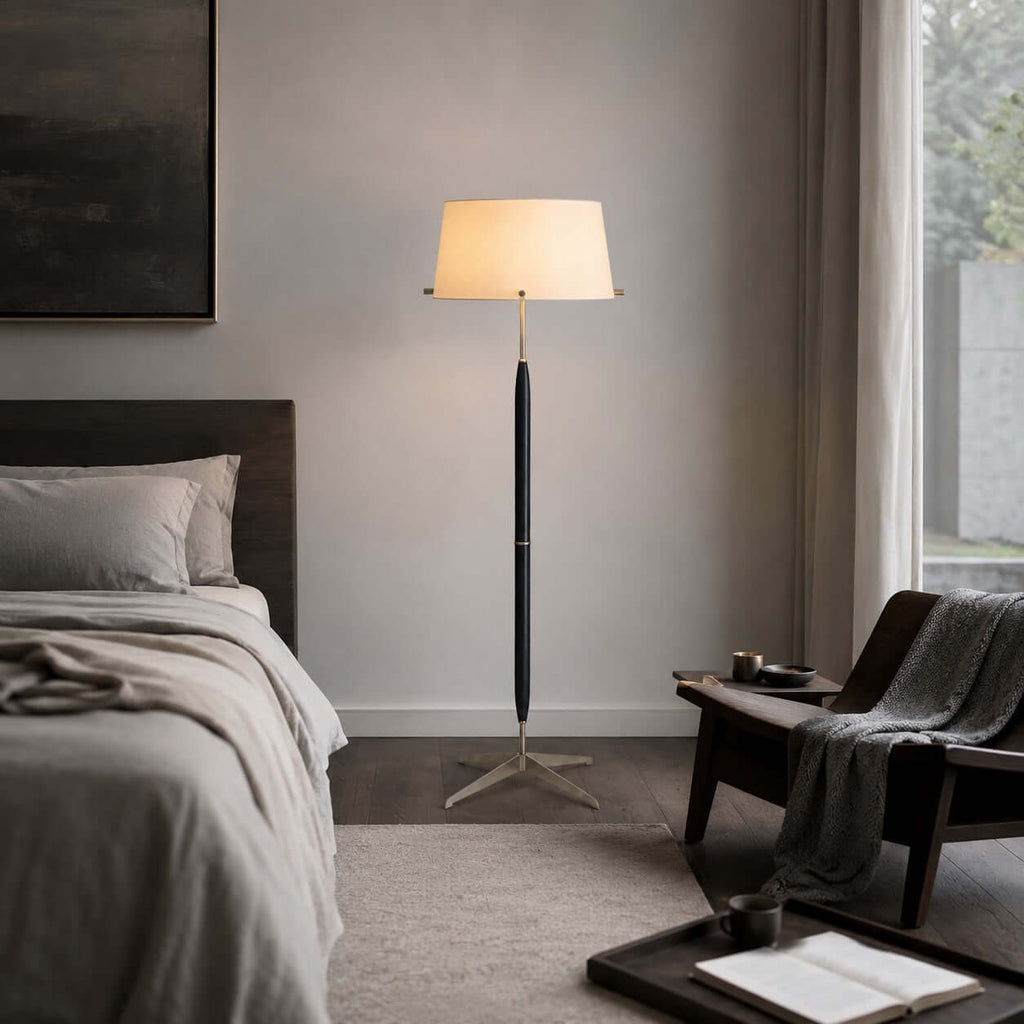 In a calm bedroom with a grey bed and a window with sheer curtains, a Clérachic floor lamp stands lit by a wooden armchair.