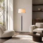 A Clérachic floor lamp stands between a curved cream armchair and a sofa, with a low round wooden table in front.