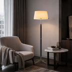 A lit Clérachic floor lamp stands by a soft pink armchair with a grey throw, with a small round marble side table next to it.