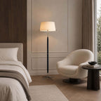 This Clérachic floor lamp stands by a bed with white linen, a cream swivel chair and a dark round side table, in front of wall panelling.