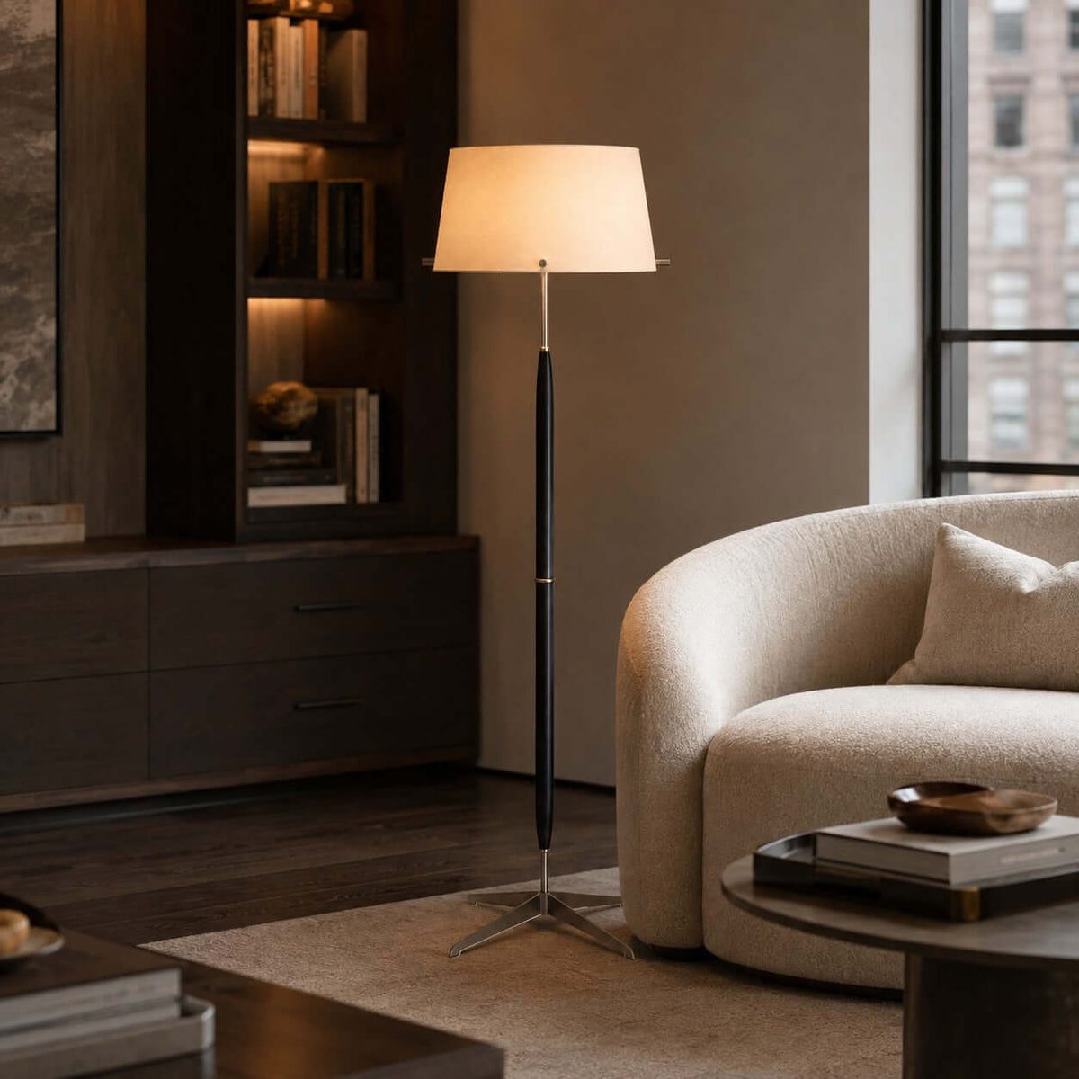 Beside a lit bookshelf and a cream bouclé armchair, a Clérachic floor lamp stands over a low round table with books.