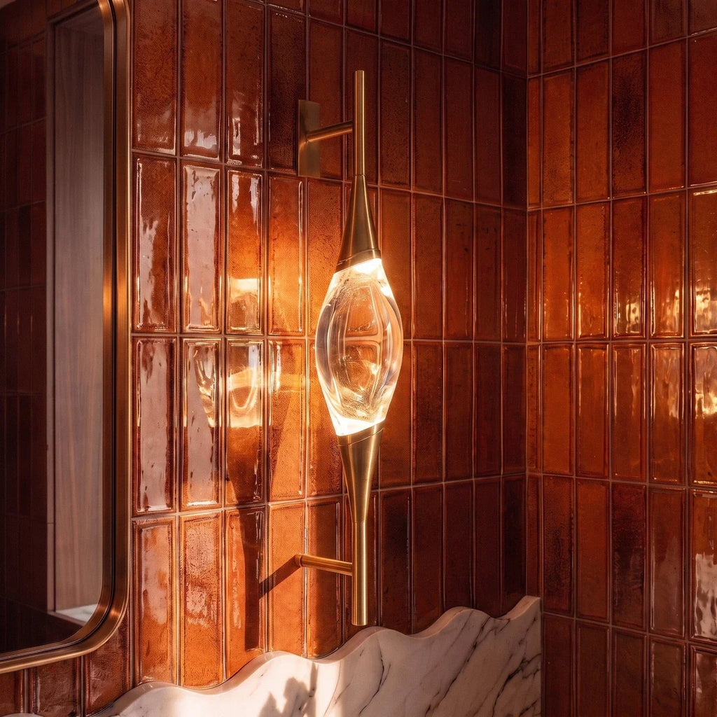 Clérachic gold wall light with a glowing crystal centrepiece, shown close up against rust-coloured glazed tiles and a brass-framed mirror.