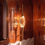 Clérachic gold wall light with a glowing crystal centrepiece, shown close up against rust-coloured glazed tiles and a brass-framed mirror.
