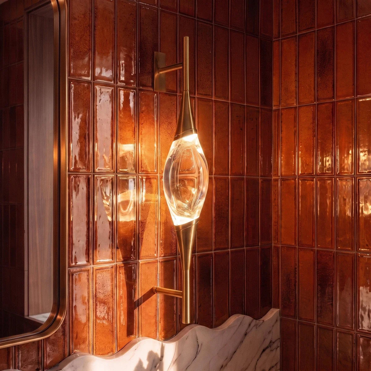 Clérachic gold wall light with a glowing crystal centrepiece, shown close up against rust-coloured glazed tiles and a brass-framed mirror.