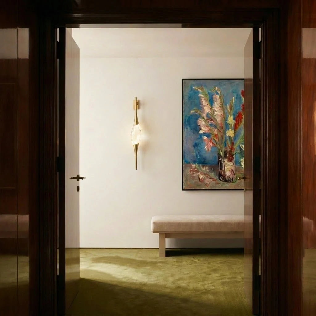 Clérachic gold wall light with a crystal centrepiece on a white hallway wall, beside a framed floral painting and a bench.