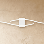 Close view of the Clérachic white wall lamp's square backplate with two wavy slim bars sweeping past each other.