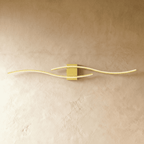 Clérachic gold wavy bar wall lamp with two slim arms crossing over a square brass-toned backplate on plaster.