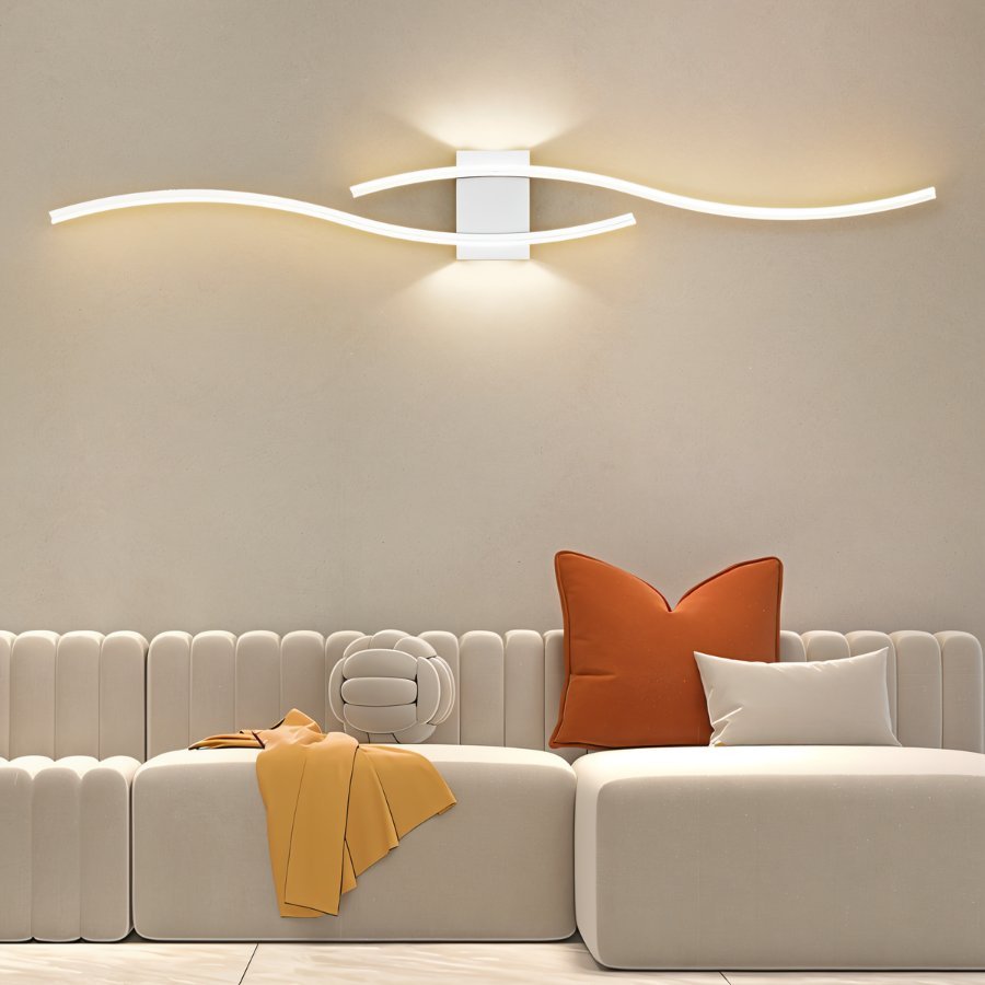 Clérachic white wavy LED wall lamp lighting a cream wall above a low modular sofa with orange and ivory cushions.