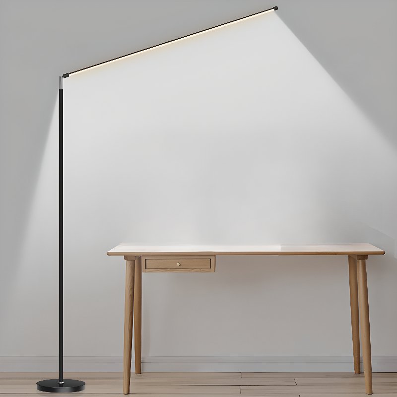 Black Clérachic floor lamp with its light bar tilted high, casting a wide glow down a grey wall beside a light wooden desk.