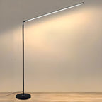 Black Clérachic floor lamp with a round base and a slim pole, its light bar lit and washing a warm glow over a beige wall.