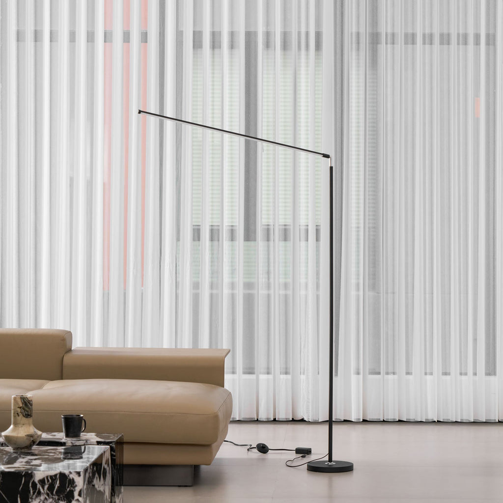 Black Clérachic slim floor lamp stands beside a tan leather sofa in front of full-length sheer curtains, with its cord on the floor.