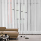 Black Clérachic slim floor lamp stands beside a tan leather sofa in front of full-length sheer curtains, with its cord on the floor.