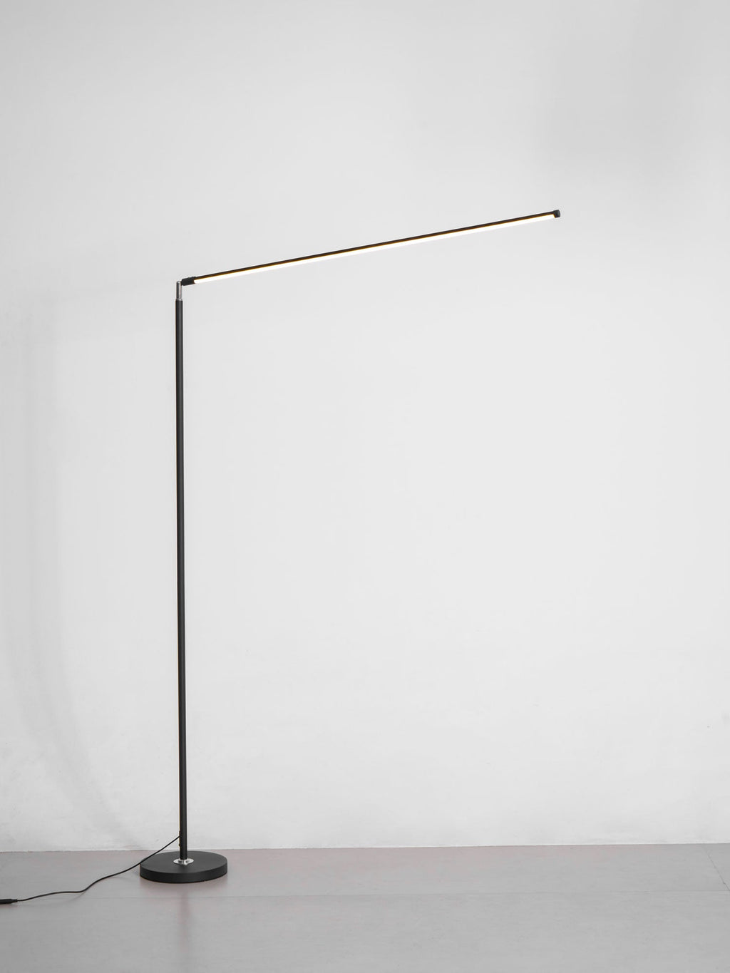 This Clérachic floor lamp stands on a grey floor with its slim pole, small round base and a long light bar lit, against a pale wall.