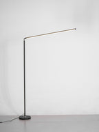 This Clérachic floor lamp stands on a grey floor with its slim pole, small round base and a long light bar lit, against a pale wall.