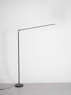 Unlit version of this Clérachic floor lamp with a black pole, a round base and a thin light bar held out at an angle, on grey.
